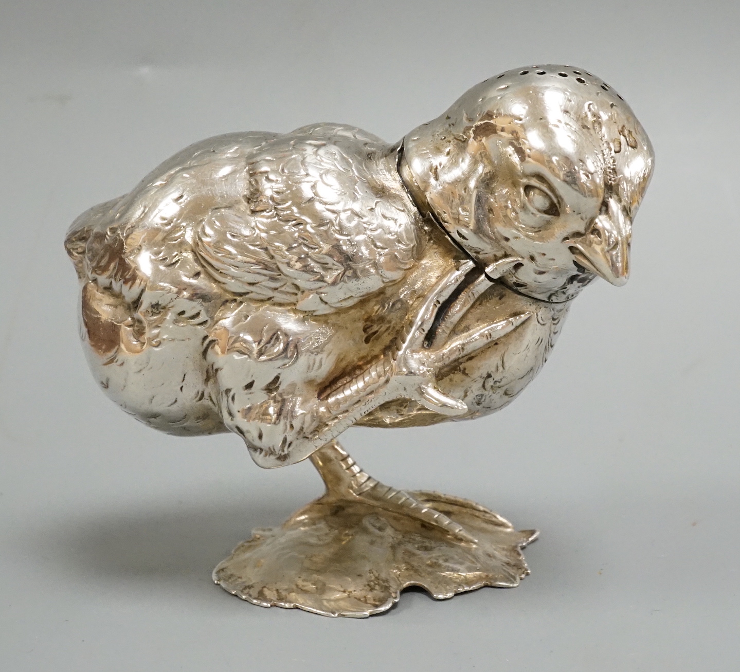A novelty white metal pepperette, modelled as a chick on one leg, scratching, height 89mm.